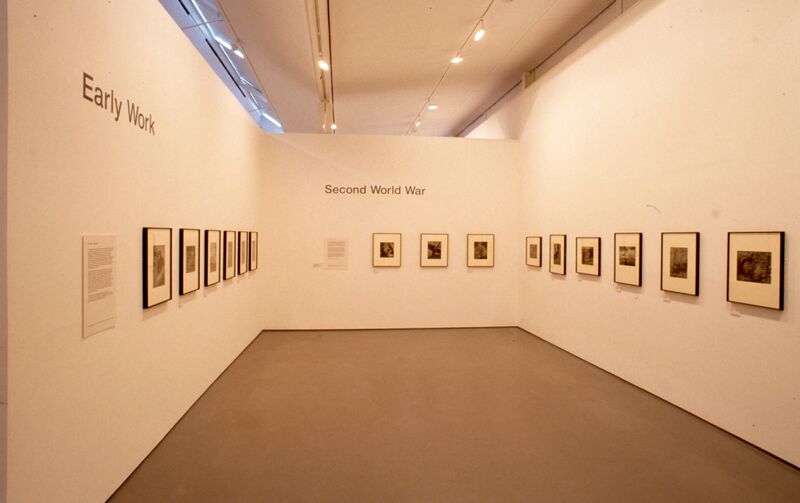 Small black-and-white photos on white backing hang on white walls. Text above the frames on one wall says 'Early work', while on the other wall the text says 'Second world war'