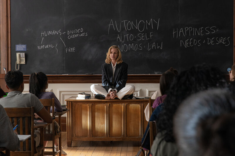 A teacher with blonde hair sits cross legged on her desk in front of her students and behind a blackboard.