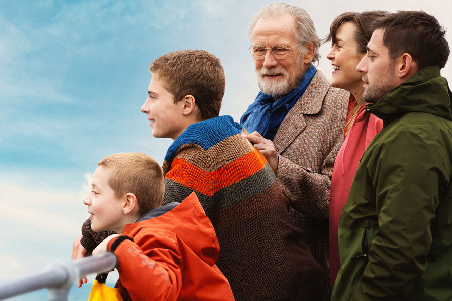 A group of five people in outdoor clothing stand together beside a railing, looking out toward a coastal view beneath a bright sky.
