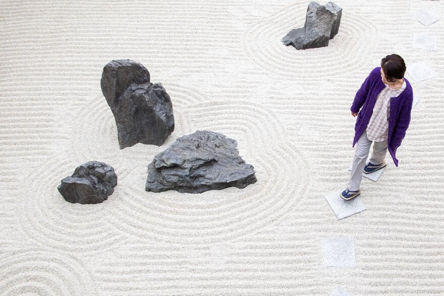 A person walks on tidy looking sand with lines in it. There are slate rocks nearby.