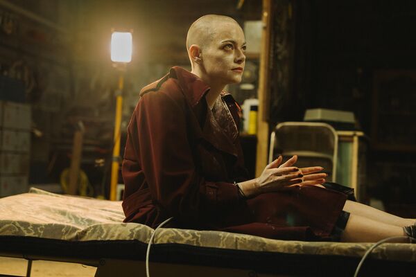 A woman with white cream on her face and shaved head sits on a camping bed in a basement.
