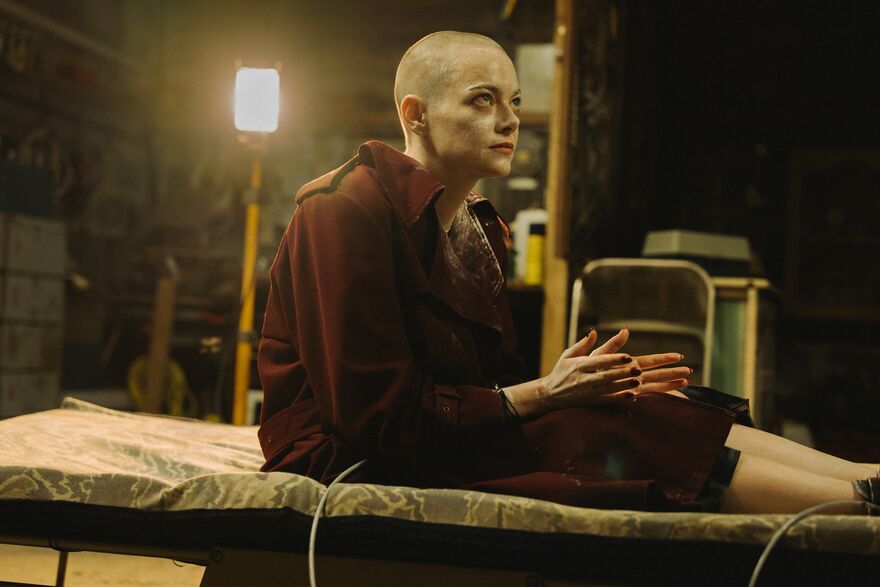 A woman with white cream on her face and shaved head sits on a camping bed in a basement.