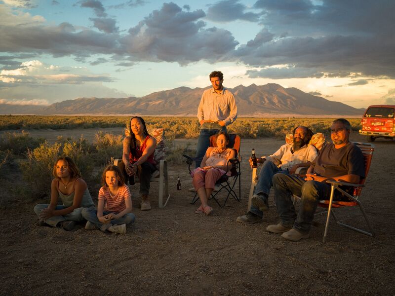 A group of people sit in camping chairs with mountains in the distance behind them and the sun setting on their faces