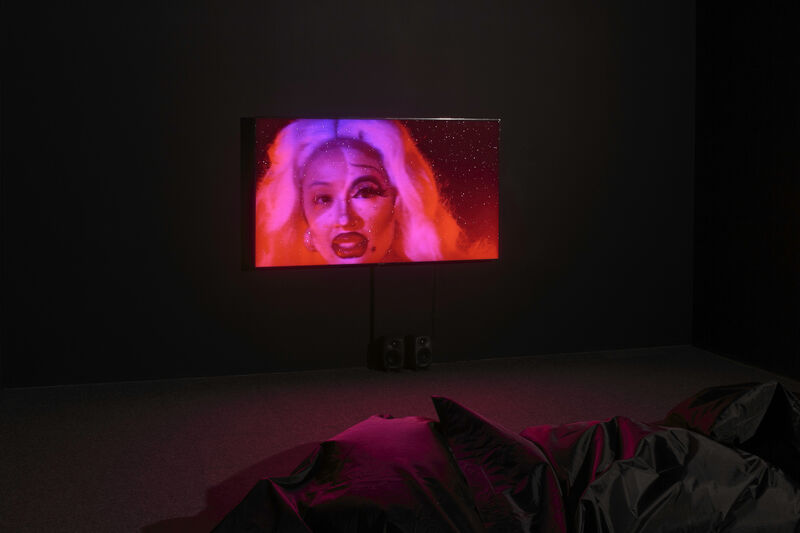 A film plays in a dark room, and we can see in the foreground, lit by the light of the screen, some beanbags on the floor. The light in the room is pink, illuminated by the vivid pink screen. The film still shows a feminine person's face, with drag-like make up over most of their face, apart from their right eye. They have heavy eye and lip makeup, and are bathed in a pink glow. 