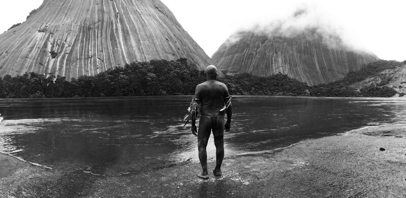 A black and white image of an amazon tribesman standing at the banks of a review looking at mountains 