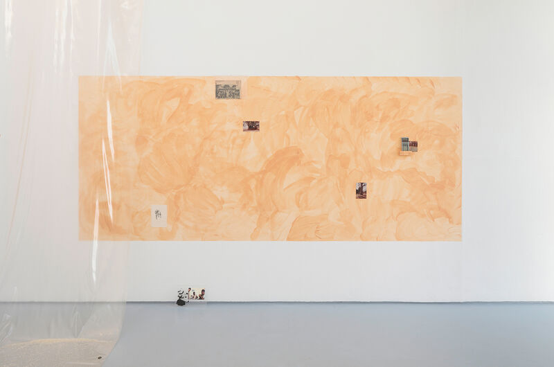 A photograph of an installation containing photographic prints incorporated onto a wall composition with an earthy orange painted ground rectangle on a white wall. To the left of the image can be seen some hanging plastic sheeting. Small objects are laid carefully on the floor under the wall based work. 