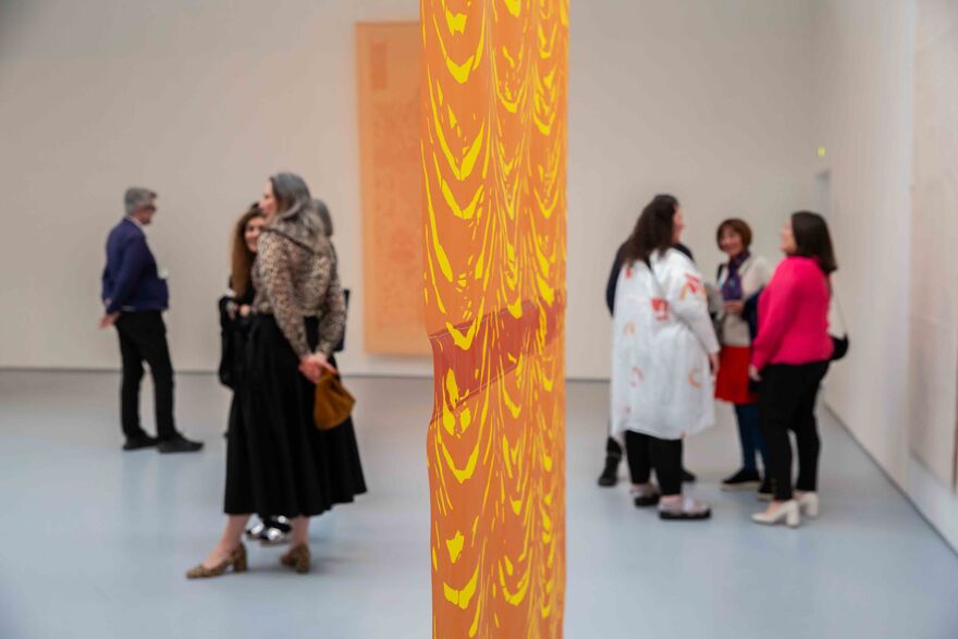 People stand in the gallery at Sukaina Kubba's exhibition opening. A yellow and orange artwork hangs in the middle of the room.