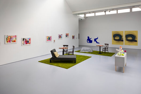 A gallery space with colourful pictures on the wall and a seating area with some sculptures on a small plinth 