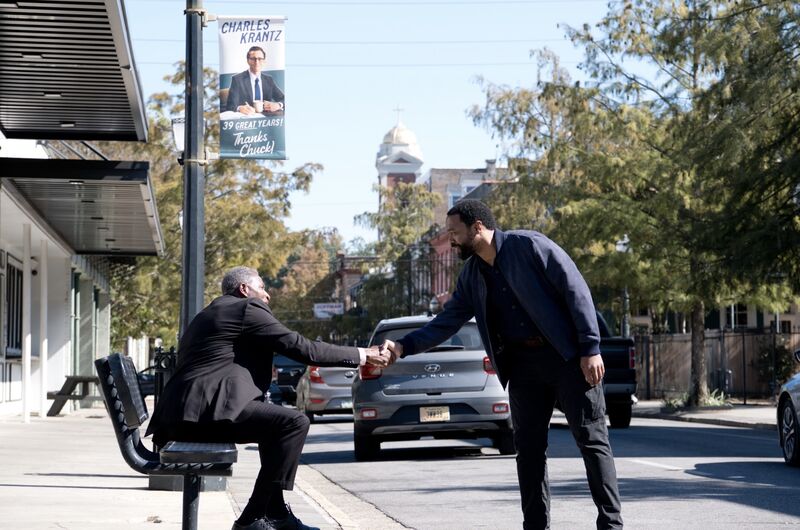 A man sitting on a park bench shakes hands with another man standing on the road.