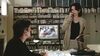 A man and a woman sit in a room surrounded by video tapes while a television plays.