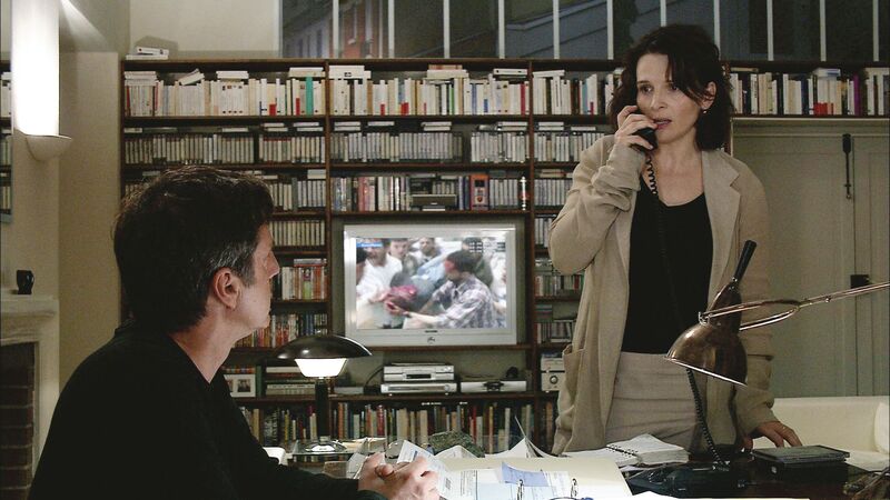 A man and a woman sit in a room surrounded by video tapes while a television plays.