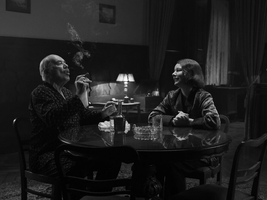 An older man and a woman at a table. He smokes a cigar, she smiles