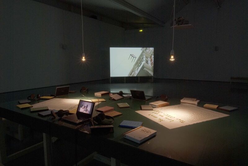 From Johanna Billing's exhibition. A large rectangular table is covered in books, tapes and posters. In the background, a projector shows an image of a table flying through the air.