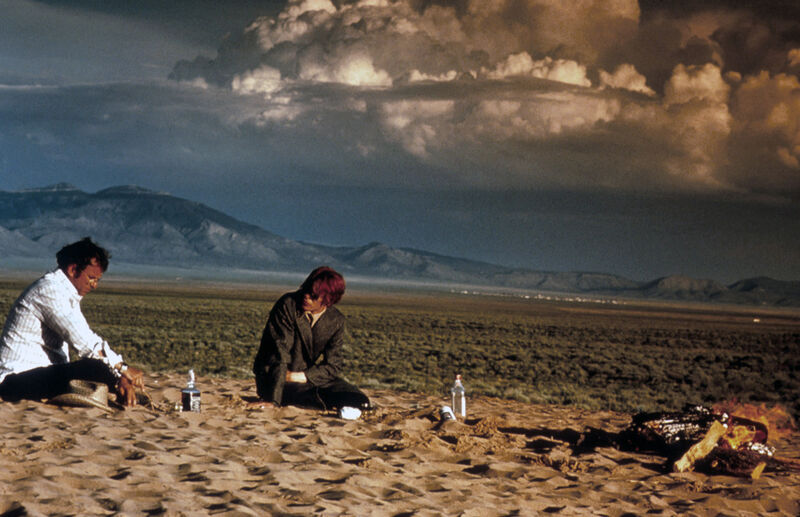 Two men sit in the desert beneath a cloudy sky
