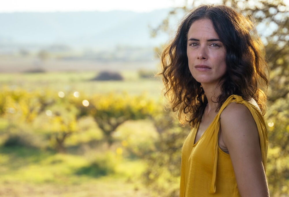 A woman with brown hair and a yellow shirt stands in a field, looking into the camera.