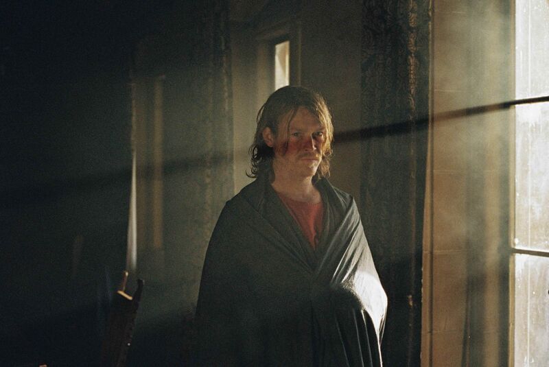 A man with a neutral expression, with a robe wrapped around him, stands in a building with the light shining in.
