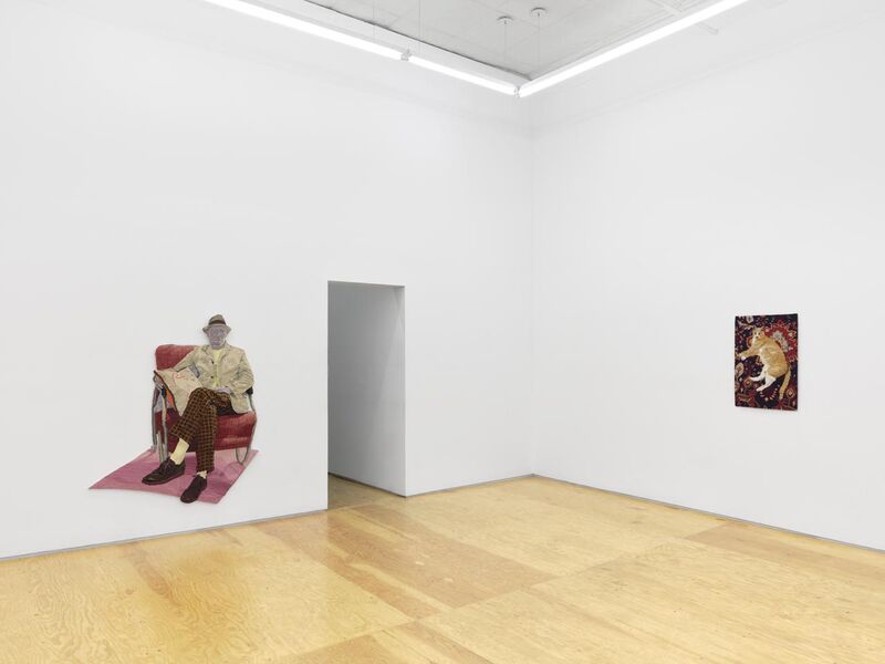 the corner of a gallery space with a tapestry of a man sitting on a chair and a cat on a rug 