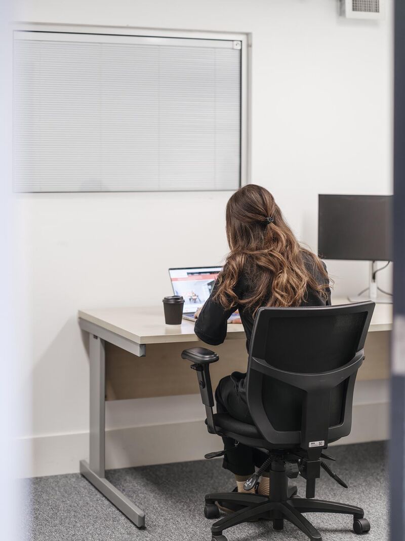 A woman with long brown hair sits at a desk with her lap top open and coffee to the side