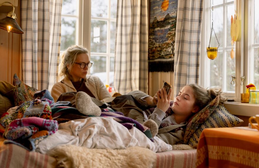 A woman and a girl sit on a bed together. The girl scrolls on her phone.