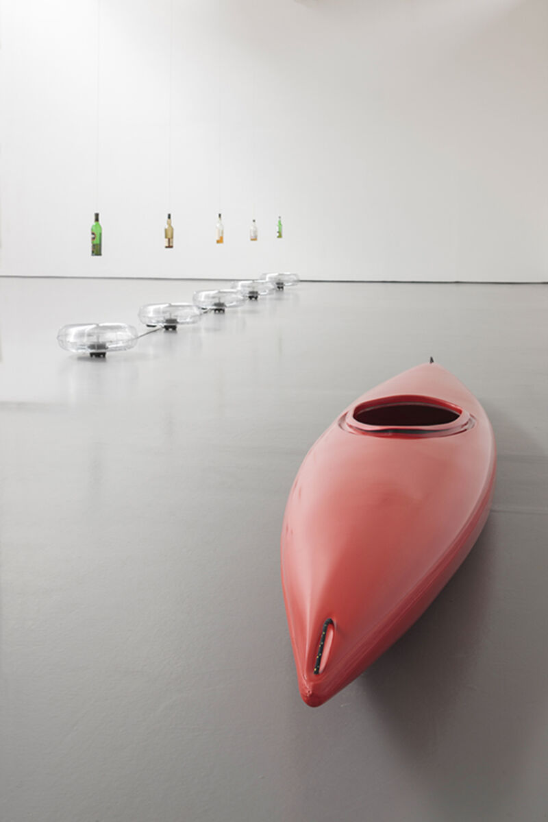 DCA Galleries during Roman Signer's exhibition. Electric fan, which are facing skyward, blow beneath glass bottles tied by string from the ceiling. Next to these, we can see a red kayak.
