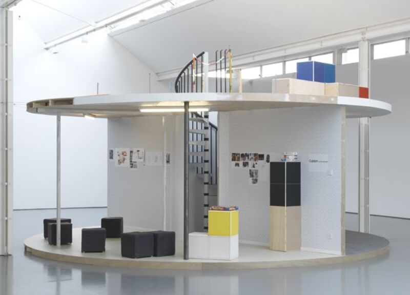 From Manfred Pernice's exhibition. A large, circular structure with two floors and a spiral staircase is segmented into different 'rooms' containing boxes and furniture.