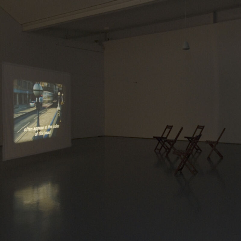 Chairs in DCA Galleries watch a video which is part of  Matthew Buckingham’s exhibition. The video shows an image of a bus driving across a bridge.