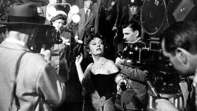 A black-and-white film still shows a glamorously dressed woman surrounded by paparazzi