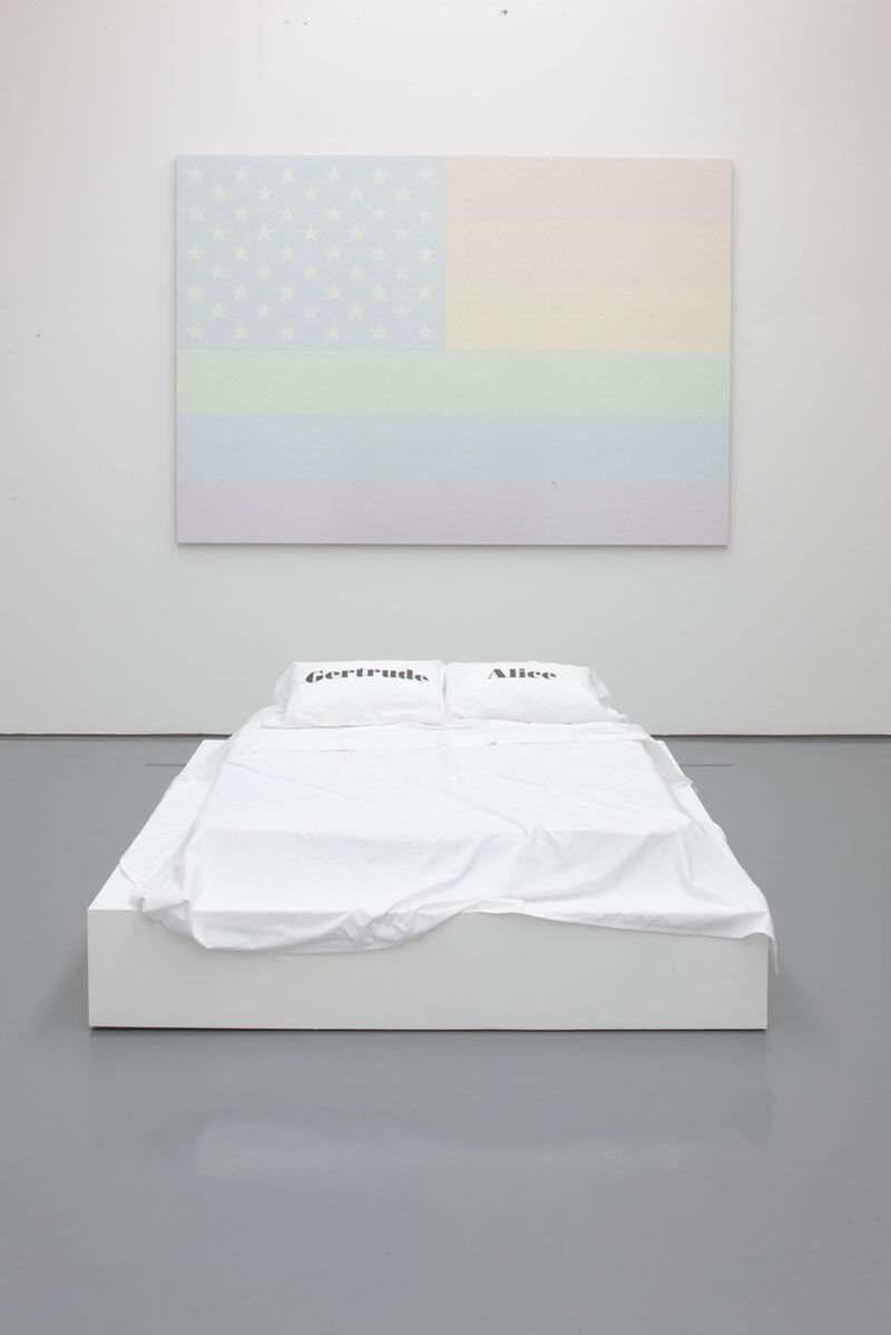 From Jonathan Horowitz's exhibition. A faded version of the USA flag, with rainbow stripes sits on the wall. In front of the flag, there is a white bed. On the pillows, one pillow has the word 'Gertrude', and the other has the word 'Alice'.