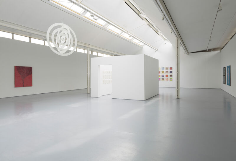 Photograph shows a wide view of gallery two from the doorway to the concourse. There is natural light, white walls and grey floor. A small boxy room has been built in the centre of the gallery, at the centre of the image. Colourful artworks are on the walls and towards the left of the image, from the ceiling hangs a white artwork made of concentric flat rings.