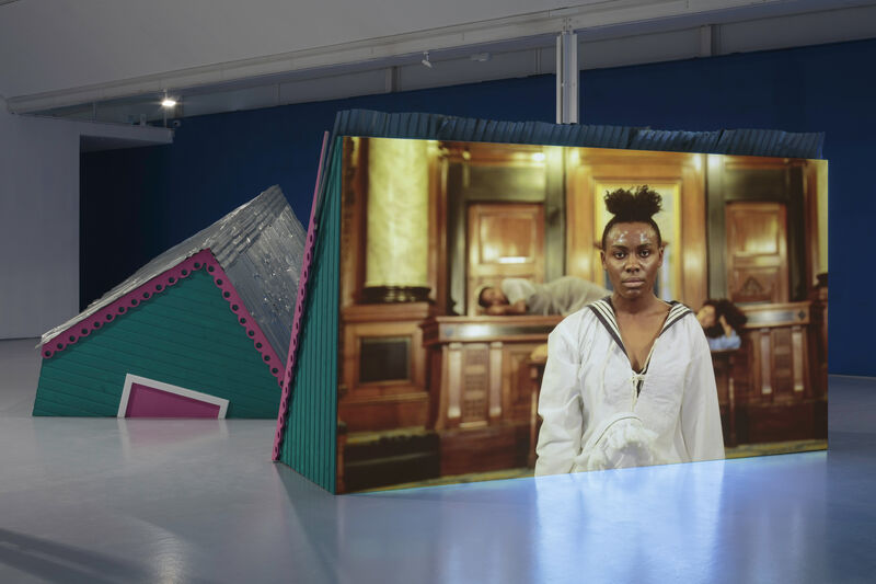 An installation image of gallery two showing a green structure like a building with a corrugated roof and a pink trim, as though it is sinking into the floor. On the back of one of these structures a film is projected. White plastic chairs are positioned in front of the screen, with iron chains around their bases. The film still a black person wearing a white shirt in a wood clad room. 