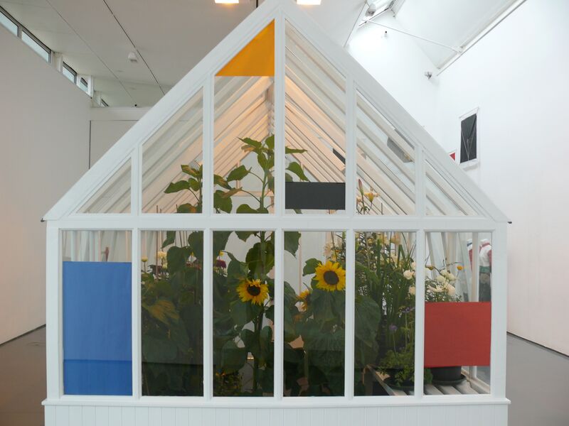 A white greenhouse sits in DCA Galleries. The greenhouse is filled with sunflowers and other flowers.