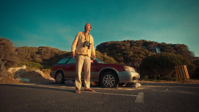 A man wearing a white shirt and binoculars stands in front of a car in a carpark.