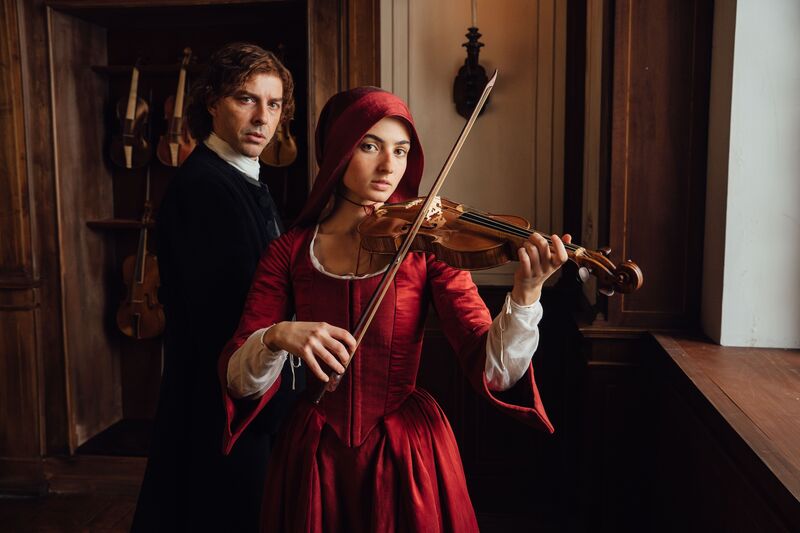 A female in the foreground wearing a period costume red in colour holding a violin with a man behind to the left in period clothing