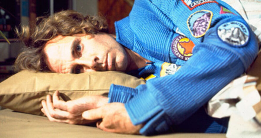 A man in a blue space suit lies on his side on a pillow with a neutral expression