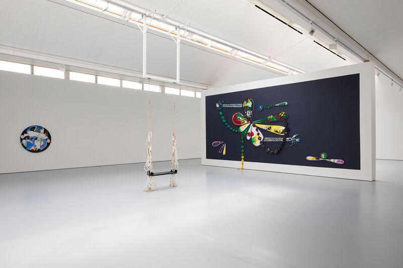 Installation documentation of Gallery 2, showing a swing hung from the ceiling towards the centre of the brightly lit gallery space. A freestanding wall towards the right hand side of the photograph shows a large mural on a dark blue background, with yellow and green elements mainly. 