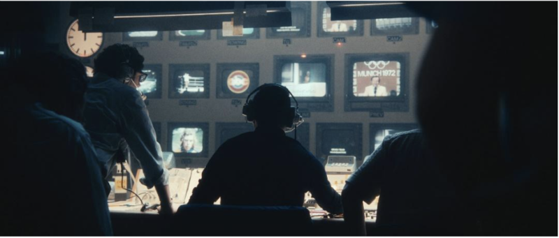 People wearing headphones sit in a broadcasting studio Iin front of rows of televisions.