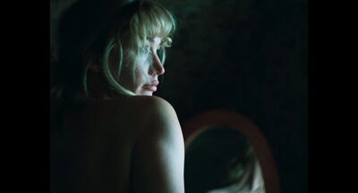 A young blonde woman sits with her back to the camera, her face and bare shoulder catching the light