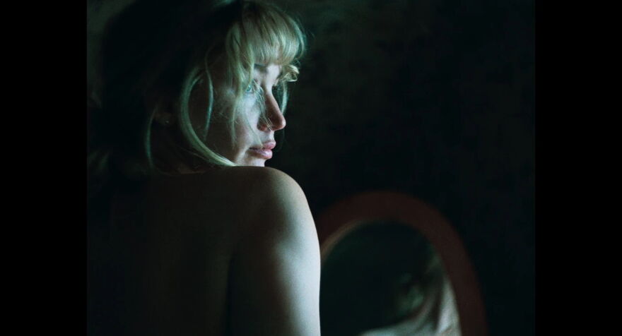 A young blonde woman sits with her back to the camera, her face and bare shoulder catching the light