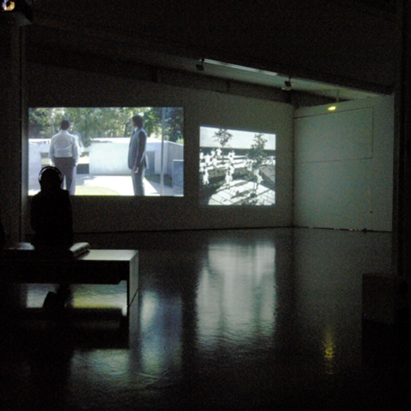 DCA Galleries during David Claerbout's exhibition. DCA Galleries are lowly lit, and a person sits watching two projector screens. 