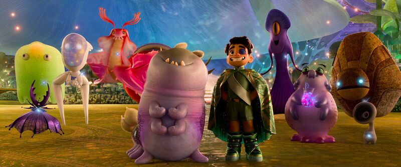 A little boy wearing a superhero costume stands smiling next to a group of unusual, colourful aliens.