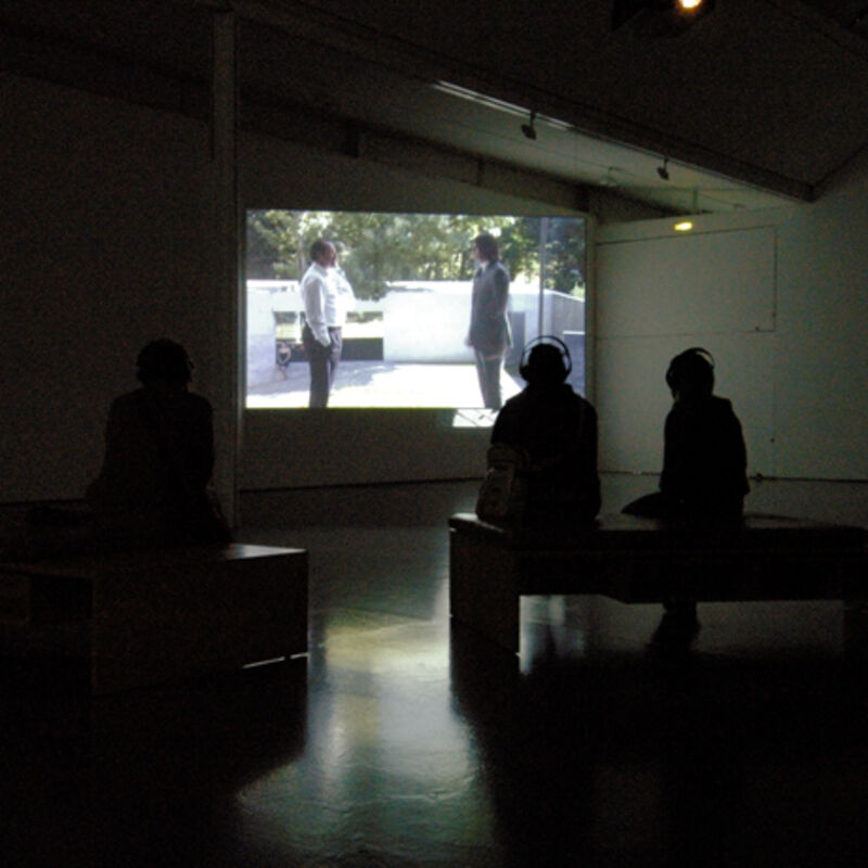 DCA Galleries during David Claerbout's exhibition - two people sit in DCA Galleries and watch two people on a projector screen.