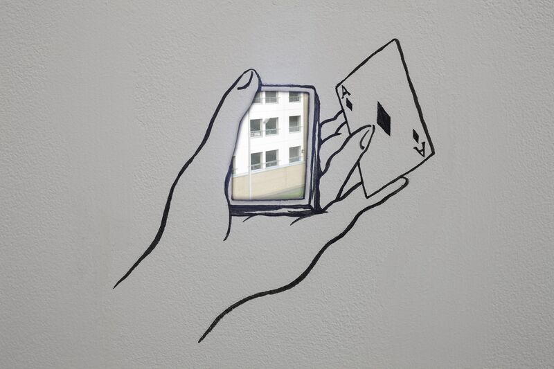 From Navid Nuur's exhibition. A mural on a white wall shows a hand holding an Ace of Diamonds between two fingers. In the rest of the hand, is a real photograph of an apartment building.