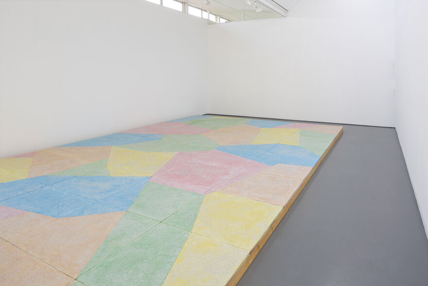 A floor based work is shown in gallery one. The walls are white and the gallery floor is painted grey. On top of this, a sculptural floor is lain in blocks, raising the floor over most of the gallery. This work is brightly coloured, with pastel blue green, yellow orange and pink, in abstract shapes.