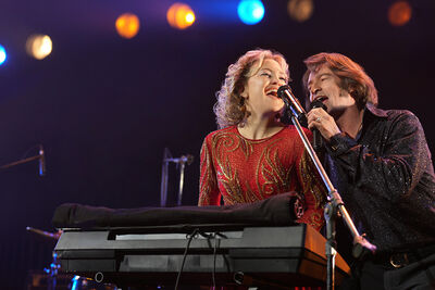 A man and a woman dressed in sparkly 70s outfits sing on stage together and play keyboard