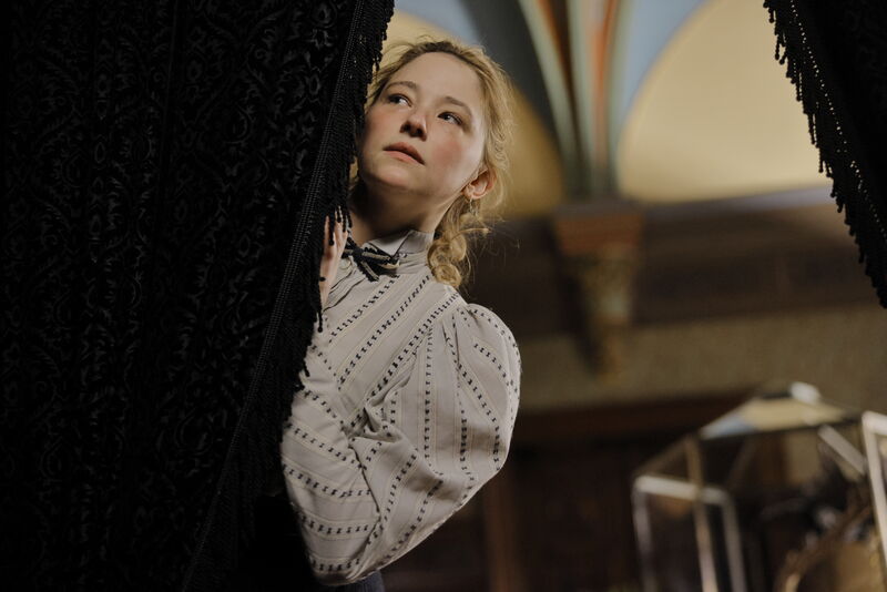 A person in a white patterned high-collar blouse peeks out from behind heavy black curtains in an ornate indoor setting, with arched architecture and warm lighting in the background.