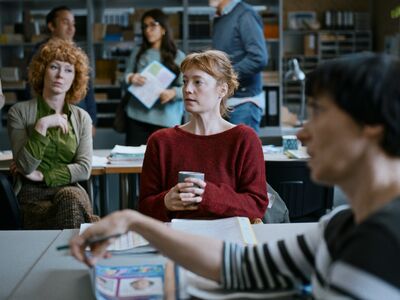 A red-headed woman sits at a desk holding a cup of coffee. Other people surround her.