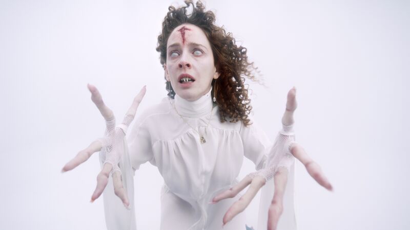 Woman reaching out with long fingers. She wears white against a white background and has a bloody cross on her forehead