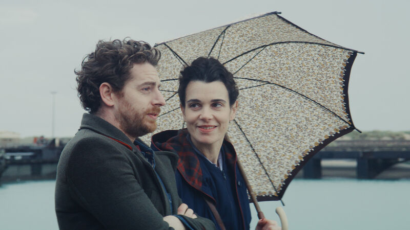 A woman holding an umbrella smiles at a man. They are walking by a pier.