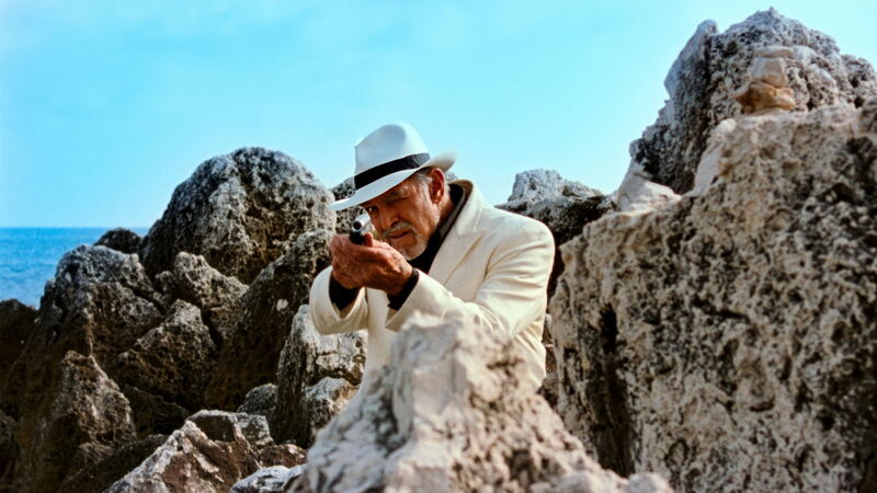 Old man in a white hat and suit holding a gun. He is surround by rocks with the sea in the background