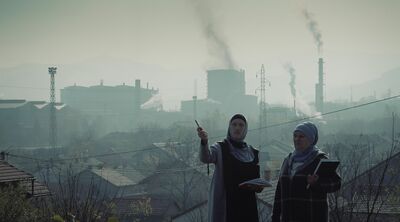 Two women stand outside in a city, while large pipes billow smoke in the background.